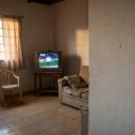 Interior de casa em Porto Velho com TV exibindo jogo do Brasil e marca de bala na parede